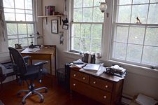 Desk and low chest of drawers covered with books in front of windows.