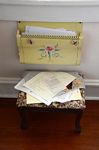 Yellow wall mounted file organizer stenciled with a rose and affixed with two bird magnets; foot stool underneath covered with disordered papers.